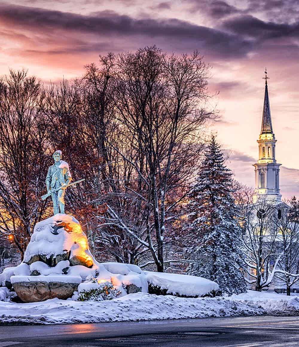 A snow-covered statue stands on a stone base near trees, with a church steeple and colorful sunset sky in the background.