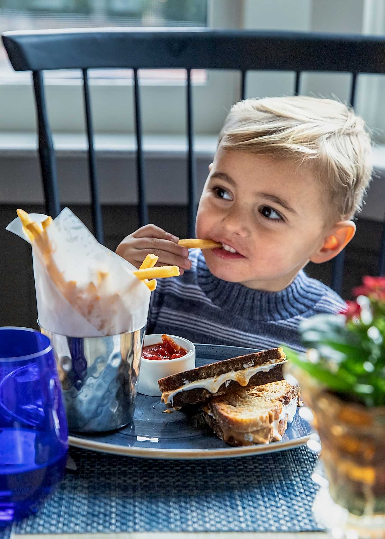 Young child with blond hair eats french fries and a grilled cheese sandwich at a table, with a small bucket of fries and ketchup in front of him.