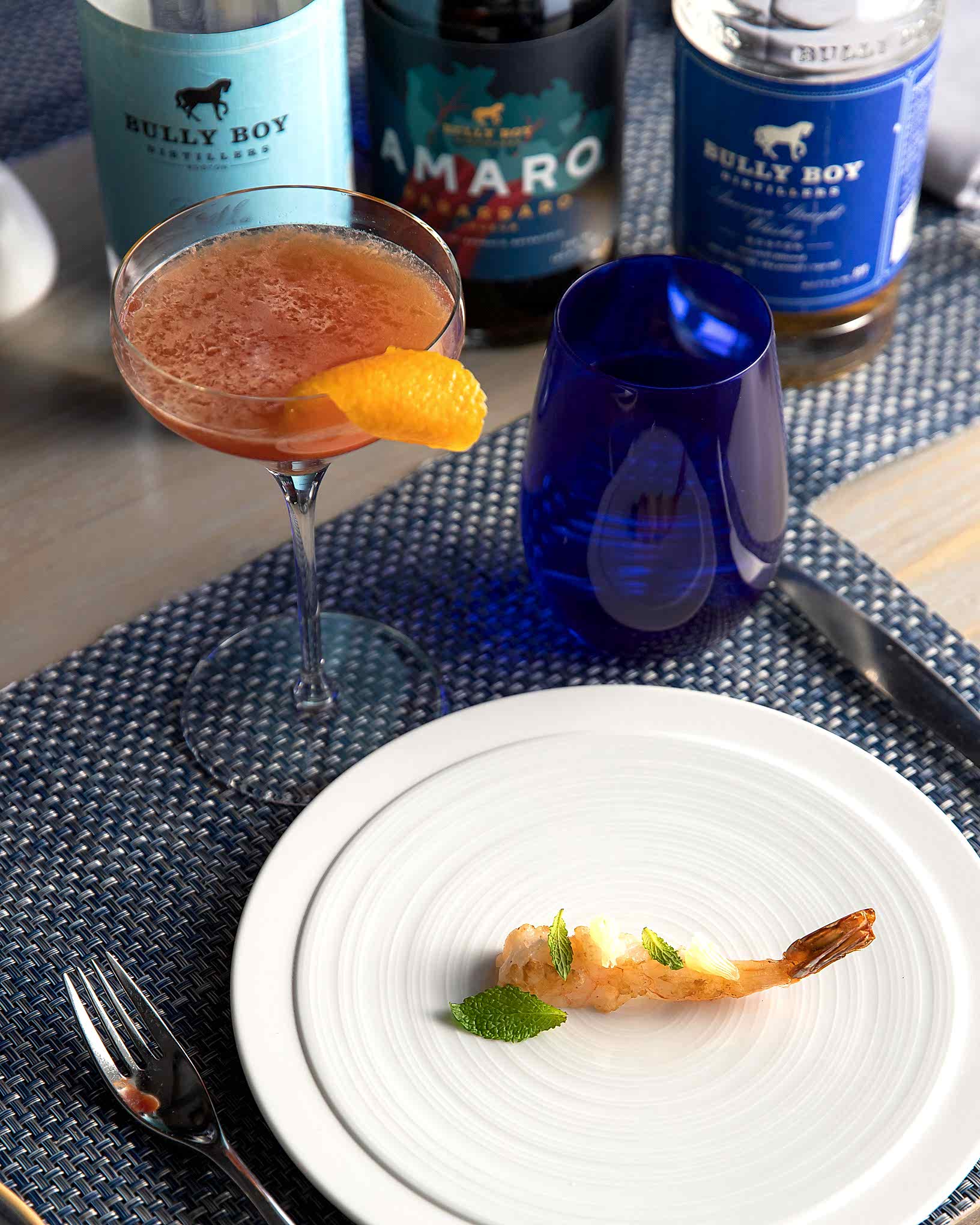 A shrimp appetizer garnished with herbs on a white plate, next to a cocktail with an orange twist and bottles on a blue placemat.