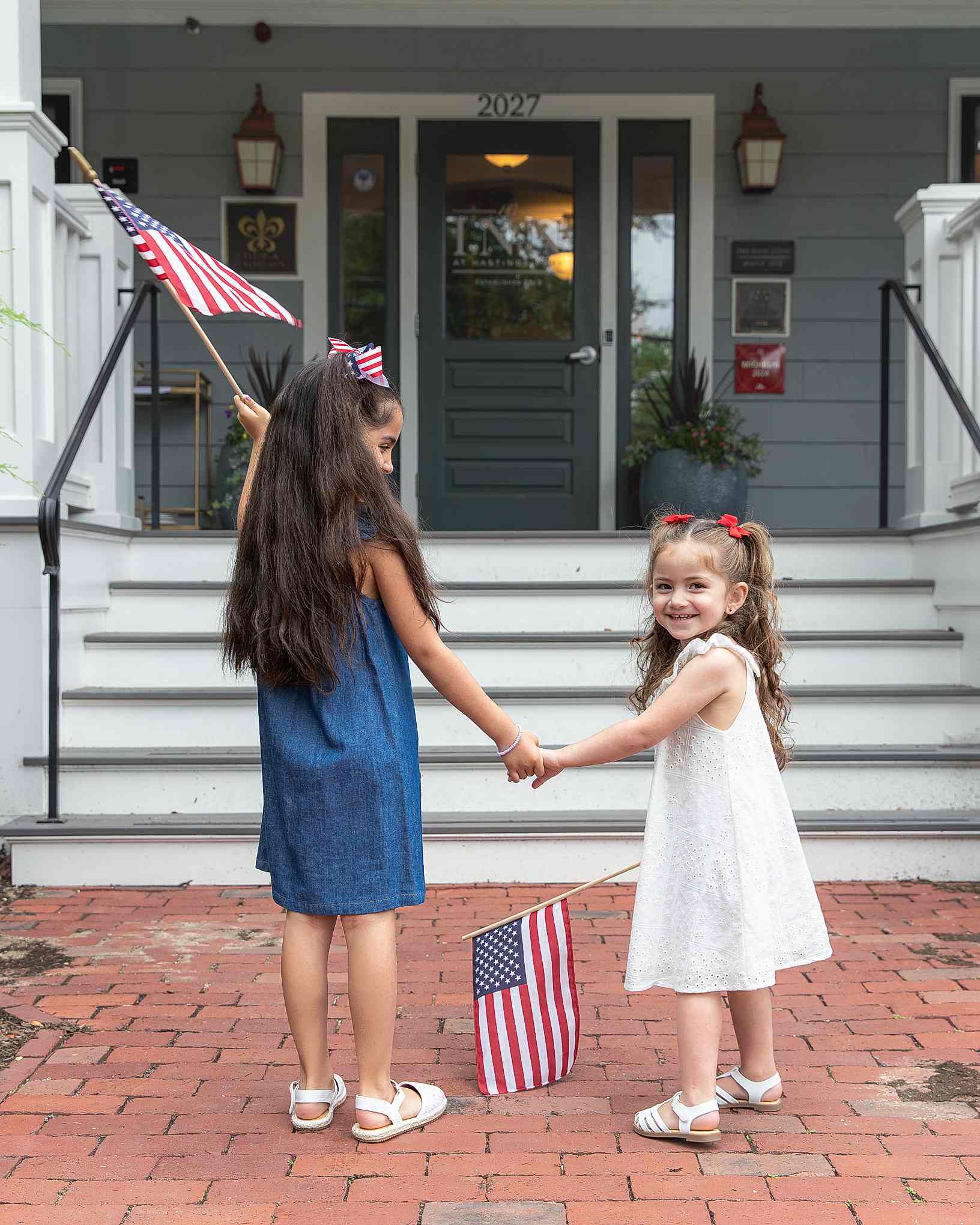 Two girls hold hands and American flags on brick pavement in front of a house.