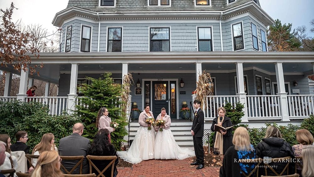 Two brides in white gowns stand together holding bouquets during an outdoor wedding ceremony in front of a large gray house, with guests seated nearby.