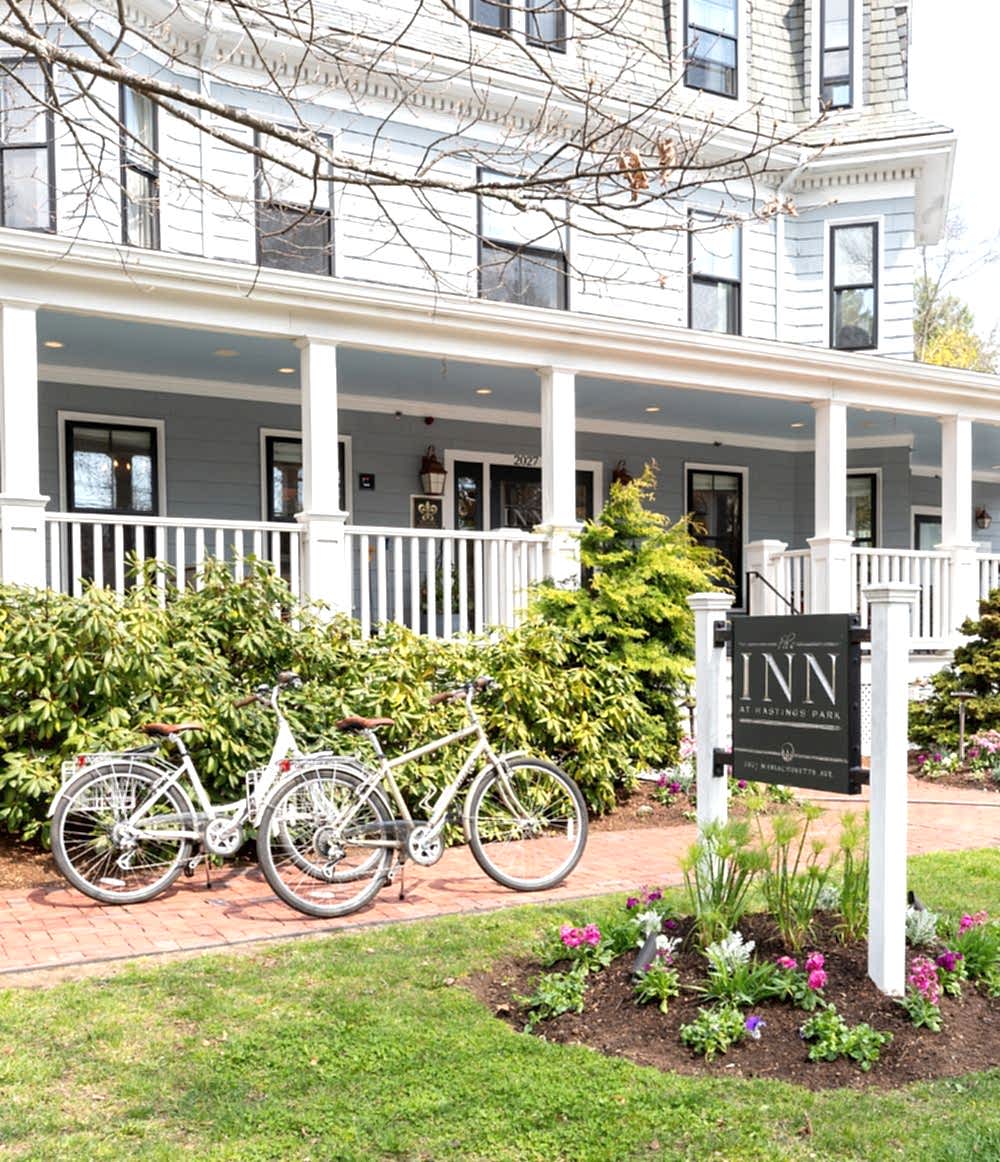 Two white bicycles are parked by a sign in front of a charming inn with a spacious porch, surrounded by greenery and blooming flowers.