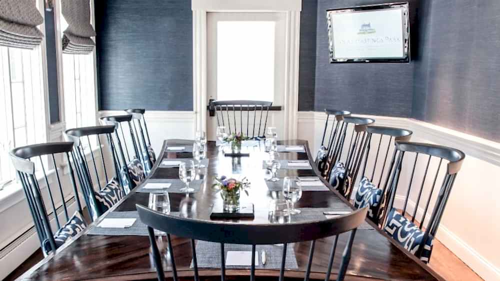 A conference room with a long table, black chairs, place settings, and a TV on the wall displaying “Inn at Hastings Park.”.