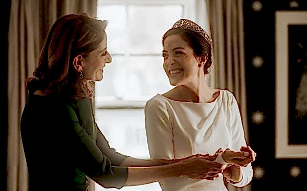 A smiling bride wearing a tiara is helped with her dress by a woman in green in a softly lit room with a window in the background.