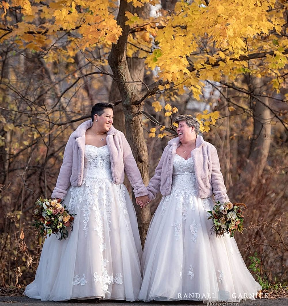 Two brides in white gowns and pink jackets hold hands and bouquets, smiling under autumn trees with golden leaves.