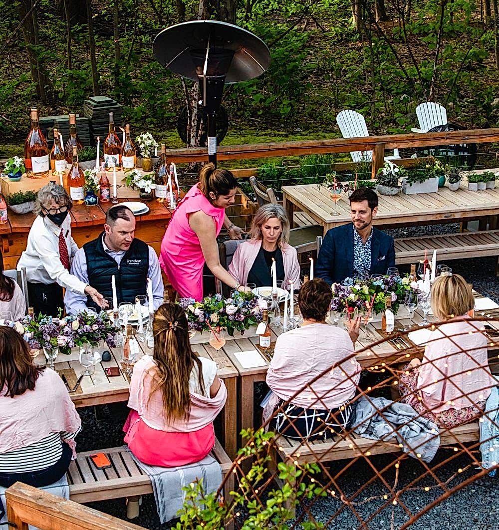 People sit at an outdoor wooden table set for a meal, surrounded by flowers and wine bottles, with servers attending to guests.