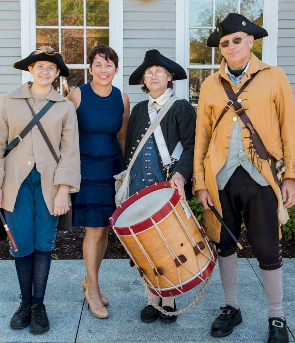 Four adults pose together outside; three wear colonial costumes, one holds a drum, and one woman in a blue dress stands between them, smiling.