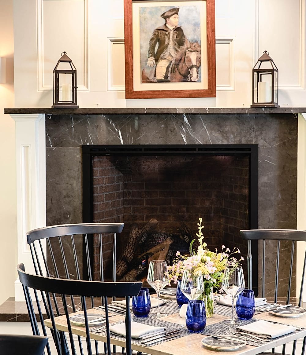 Elegant dining table with black chairs, blue glasses, and flowers set in front of a fireplace with a painting and lanterns on the mantel.
