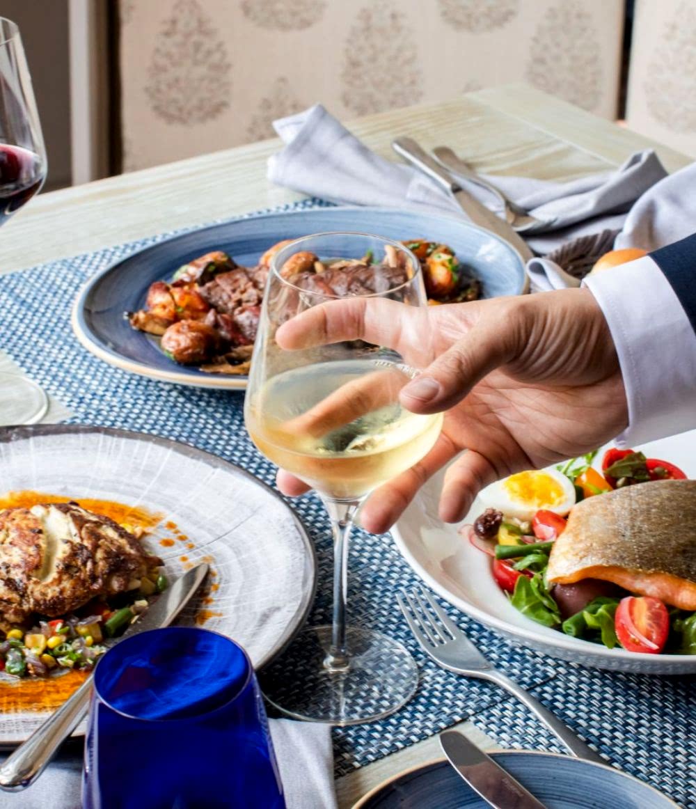 A hand holds a glass of white wine over a table set with gourmet dishes, including fish, salad, and meat, in a restaurant setting.