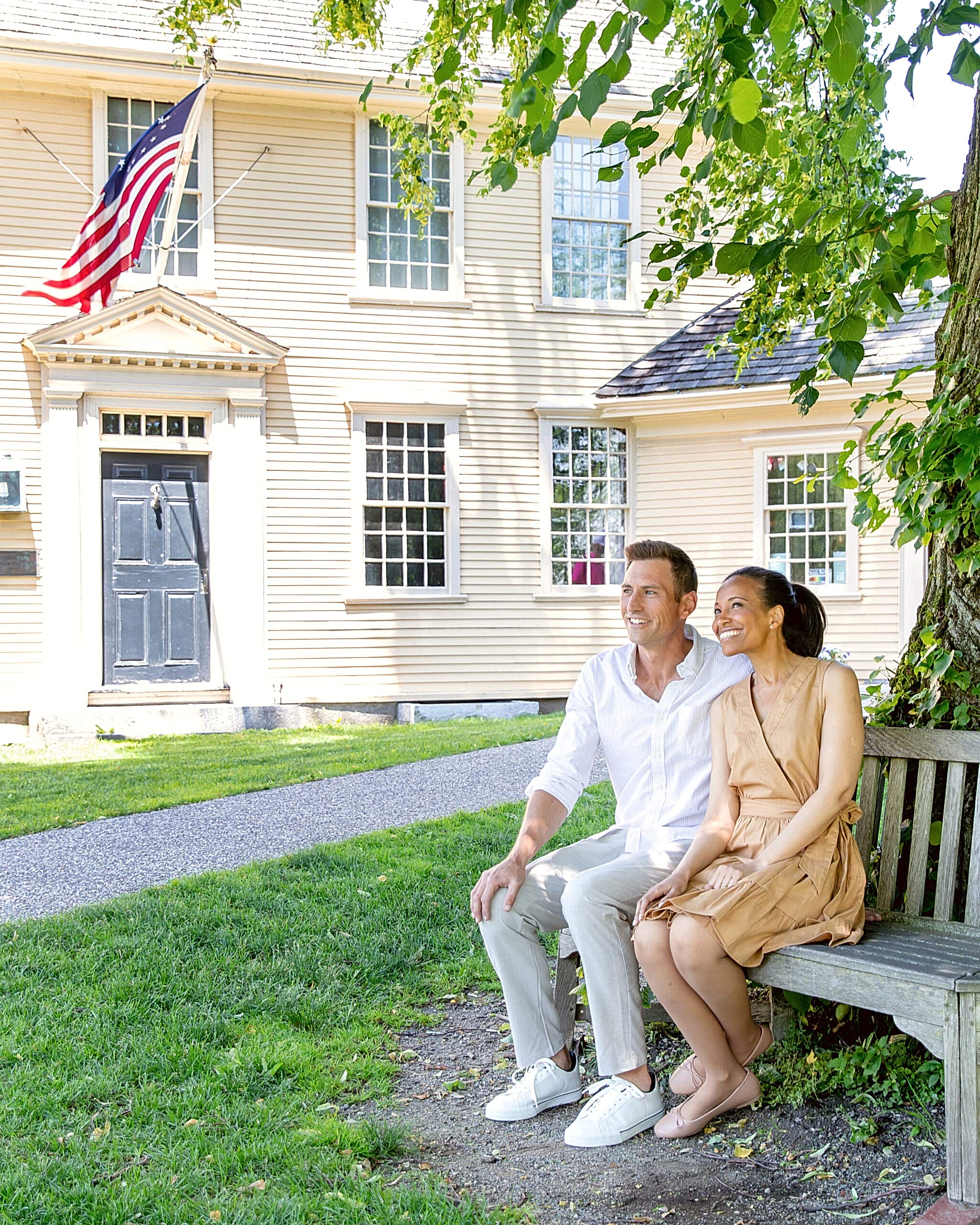 A smiling couple sits on a bench under a tree near a historic beige house with an American flag and black door, enjoying a sunny day.