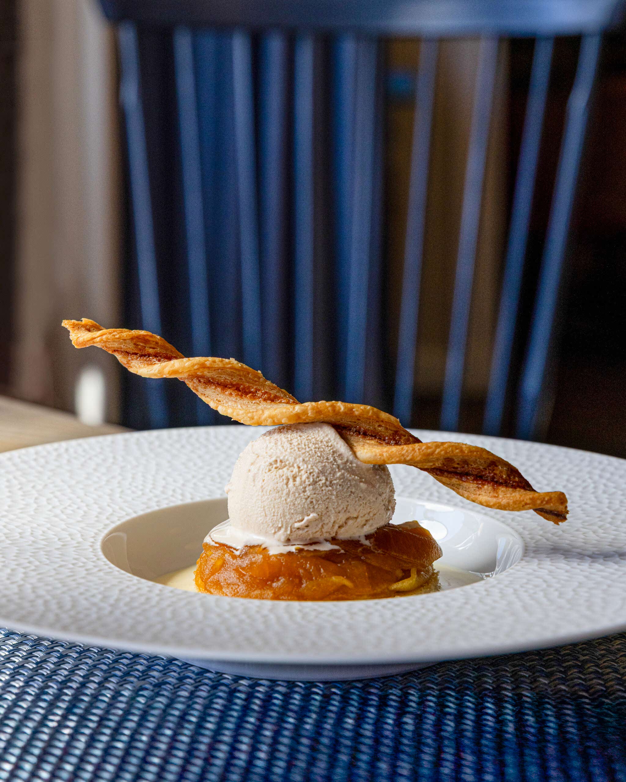 A dessert with a scoop of ice cream on caramelized fruit, topped with a twisted pastry stick, served on a white textured plate.