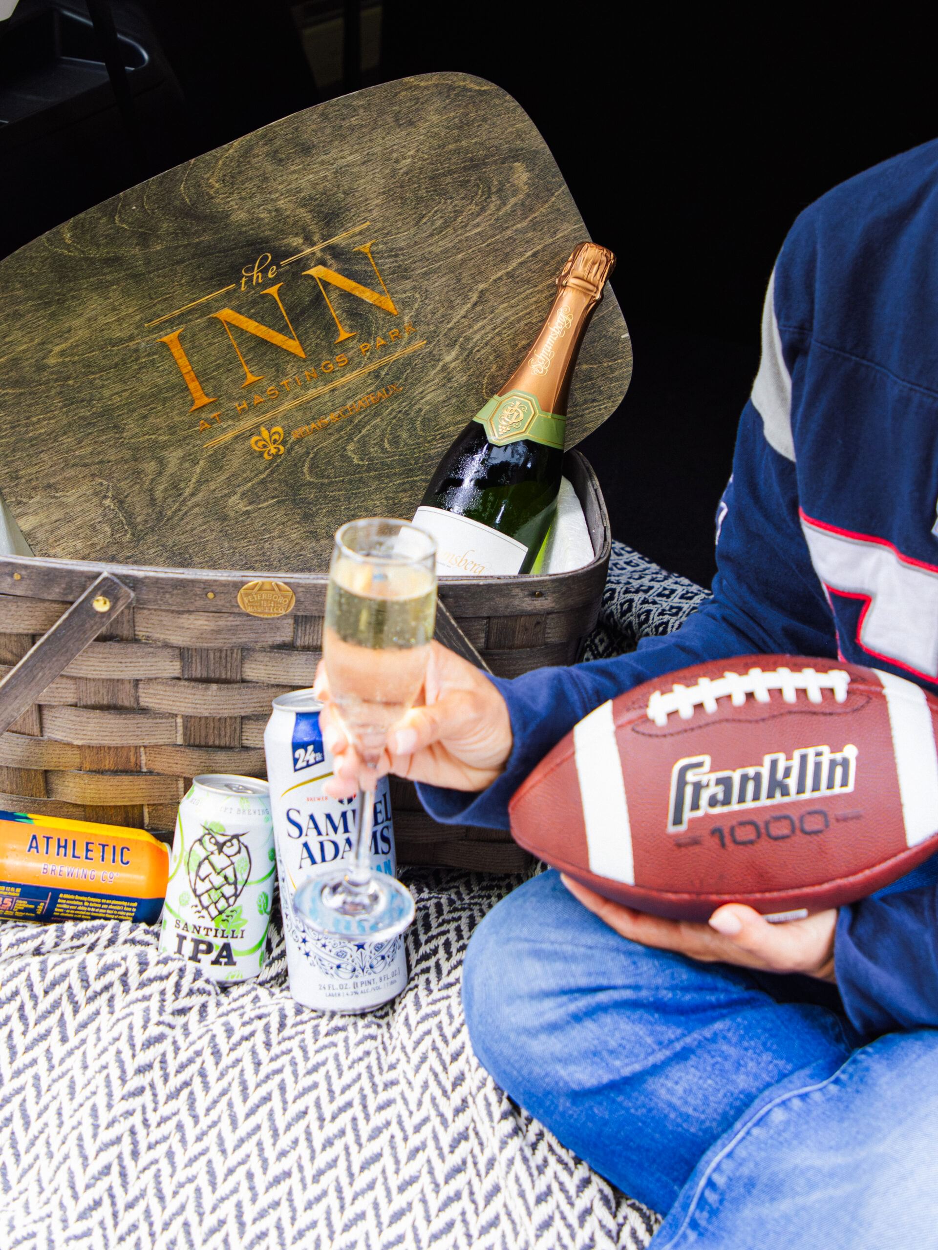 Person holding a football and champagne, seated on a picnic blanket with beer cans, a champagne bottle, and a basket labeled The Inn..