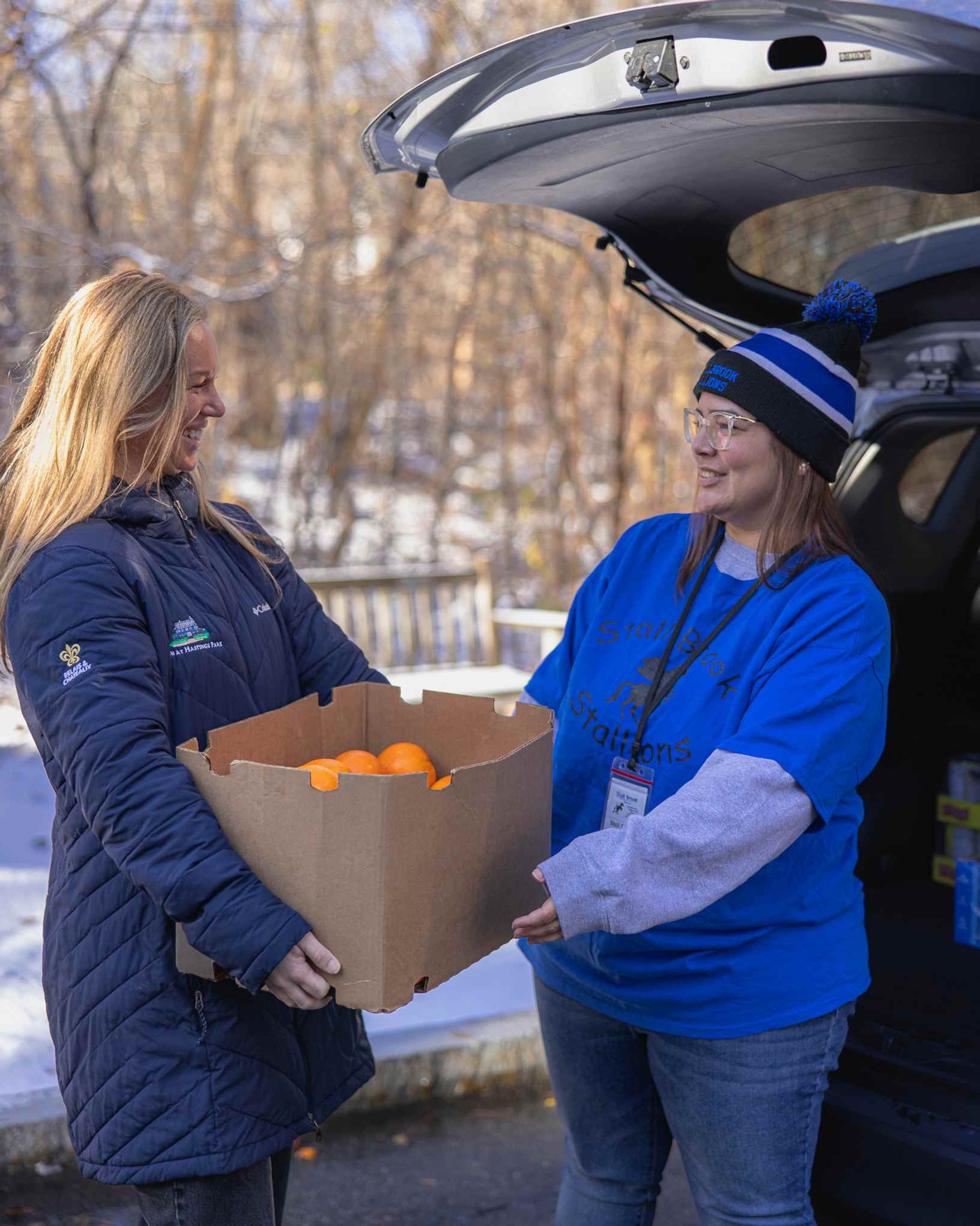 Two women smiling as one hands a box of oranges to the other by an open car trunk on a snowy day.