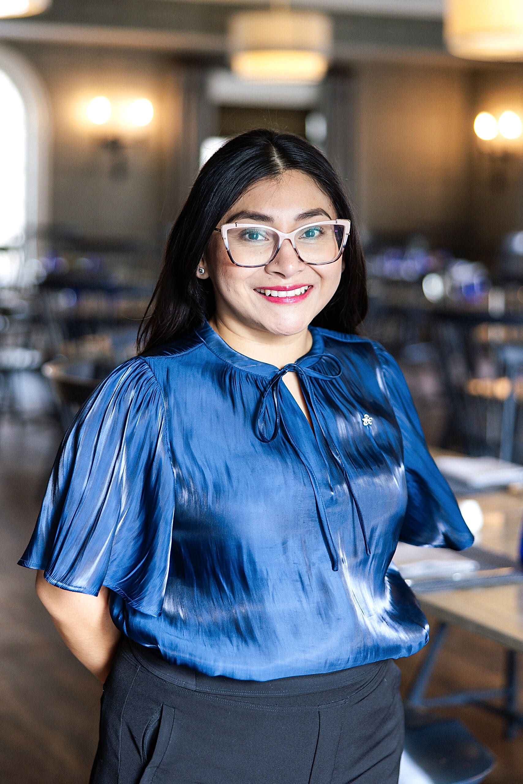 Smiling woman with long dark hair and glasses wearing a shiny blue blouse stands in a softly lit room with tables and chairs.
