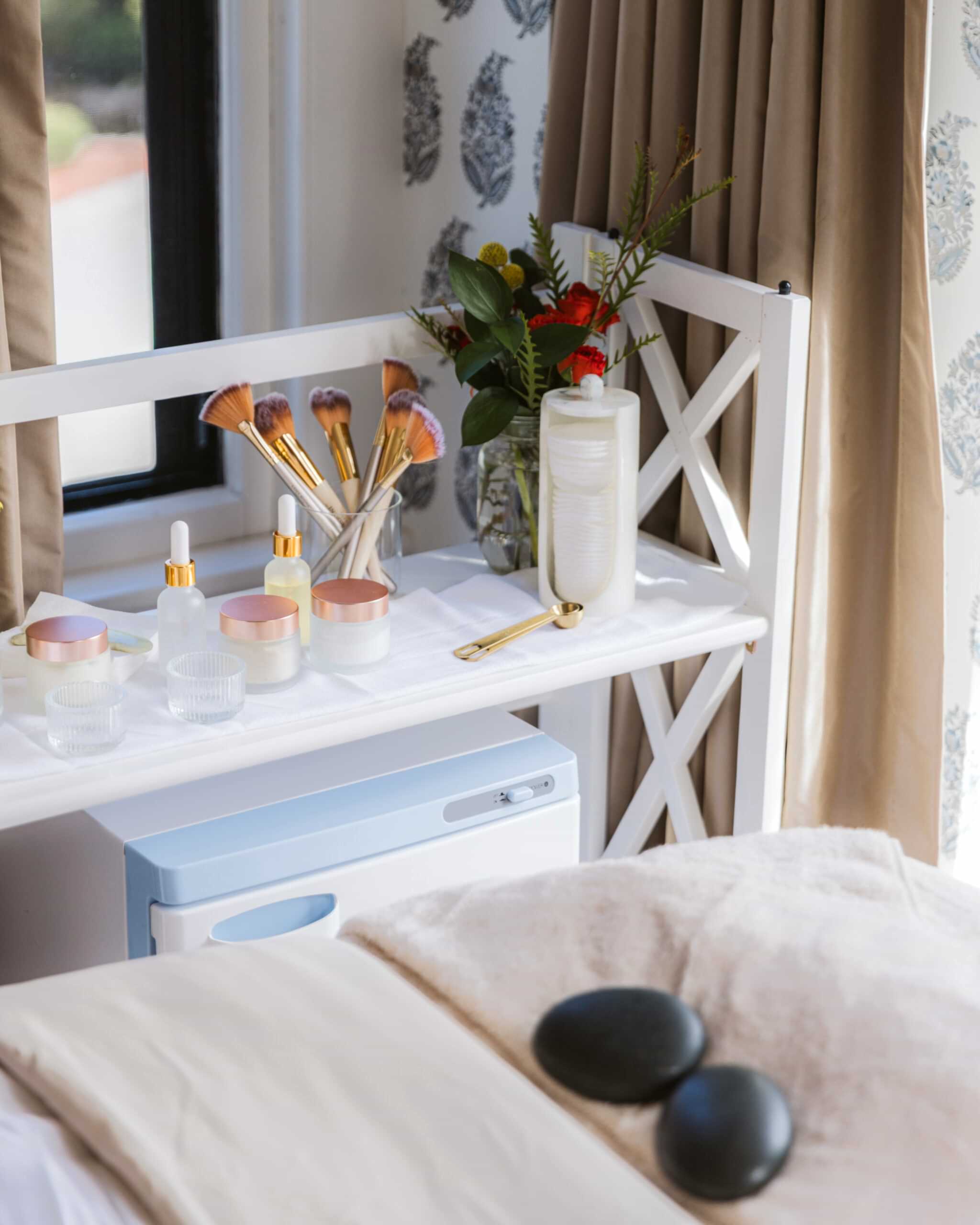 A white vanity shelf with makeup brushes, skincare bottles, and a vase of flowers next to a window. Two stones rest on a beige bed in the foreground.