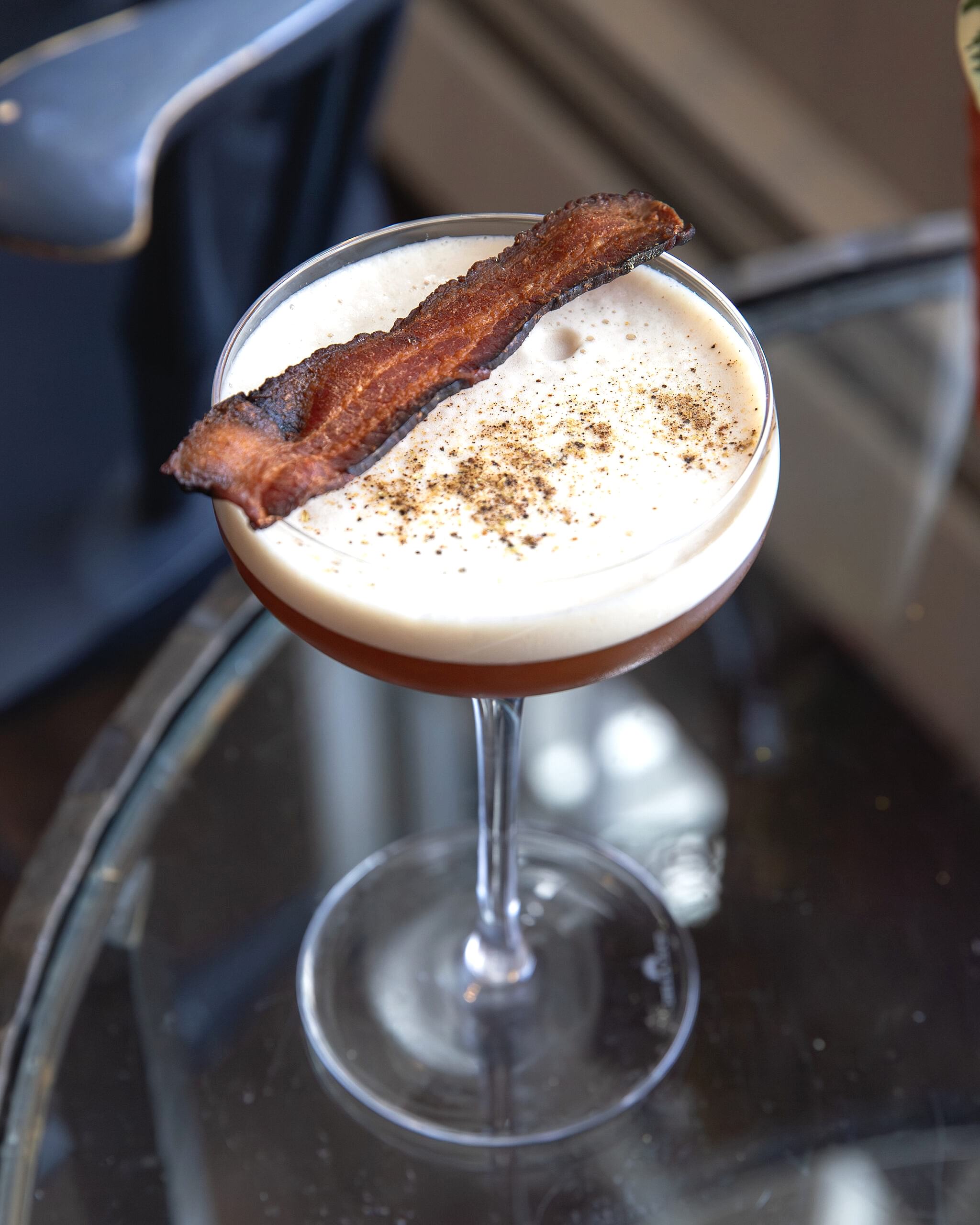 A cocktail in a coupe glass with a foamy top, garnished with a strip of crispy bacon and a sprinkle of black pepper, on a glass table.