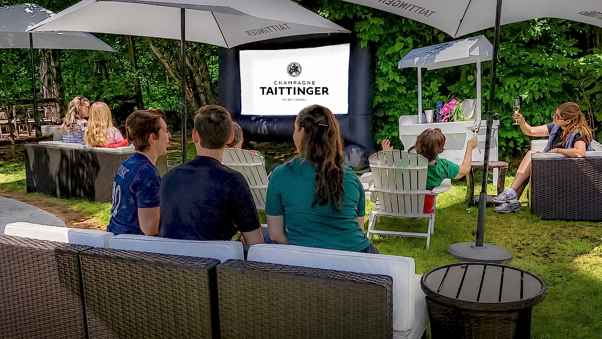 People watch an outdoor screen under umbrellas, surrounded by greenery. The screen displays Champagne Taittinger.