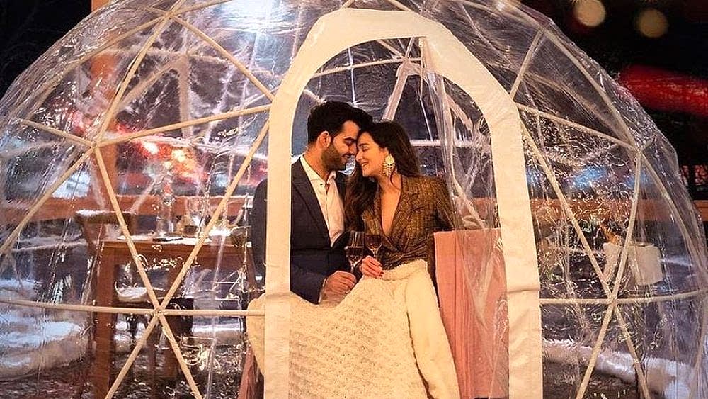 A couple sits close together inside a clear dome, smiling and holding drinks, wrapped in a blanket on a cozy evening.