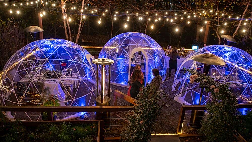 Three clear, dome-shaped dining pods glowing with blue lights under string lights at night, with people dining inside and outside the pods.