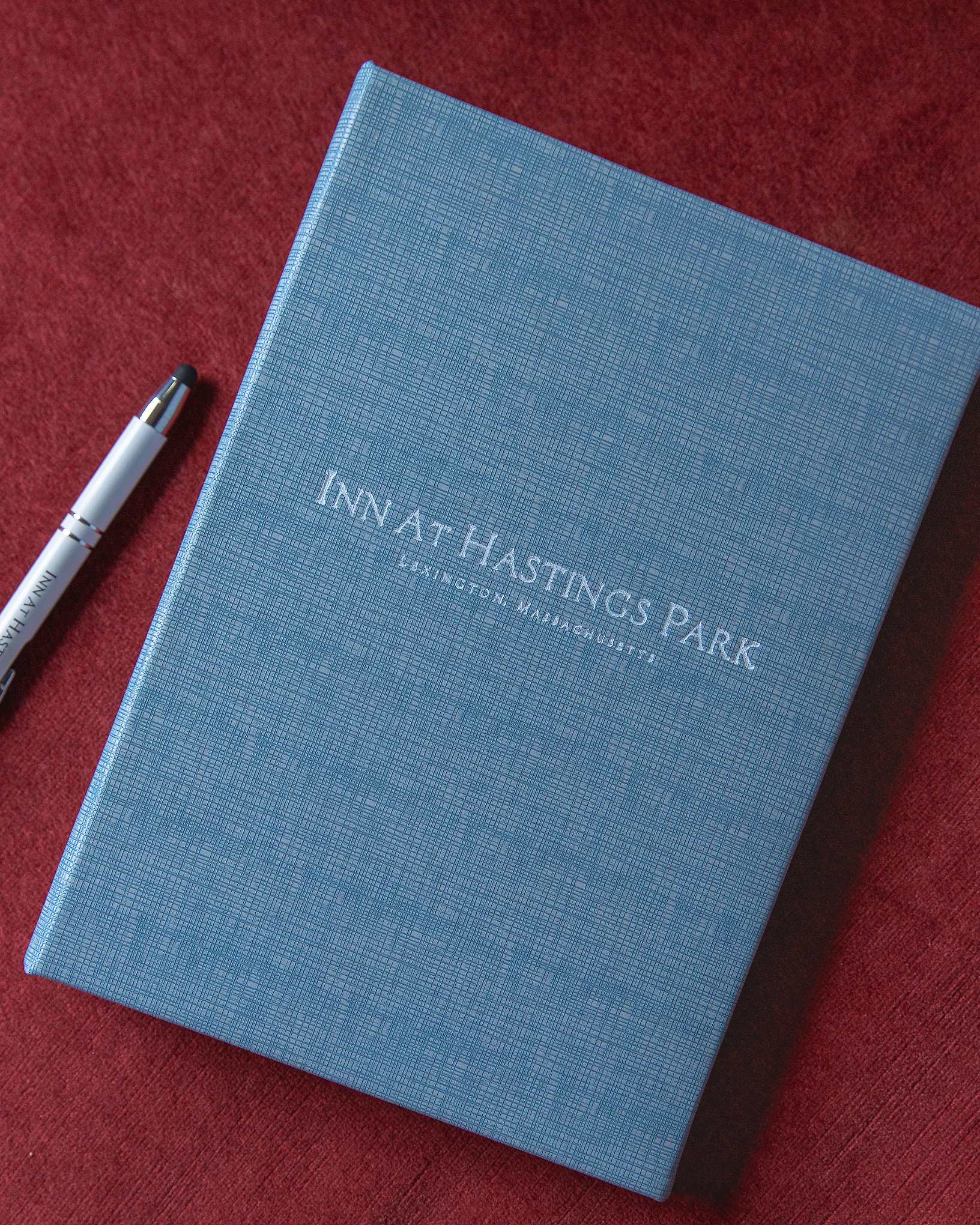 A blue textured notebook labeled Inn at Hastings Park Lexington, Massachusetts lies on a red surface next to a pen.