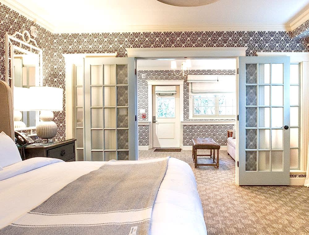 Bright hotel room with a large bed, patterned wallpaper, glass double doors, and a sitting area with a bench and windows in the background.