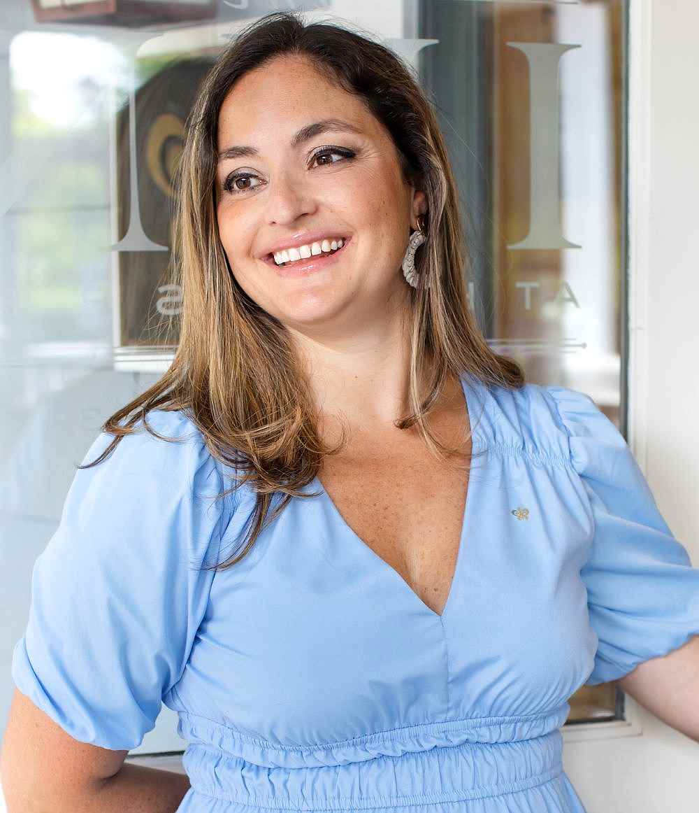 A woman with long brown hair wearing a light blue dress smiles while standing indoors in front of a glass door.