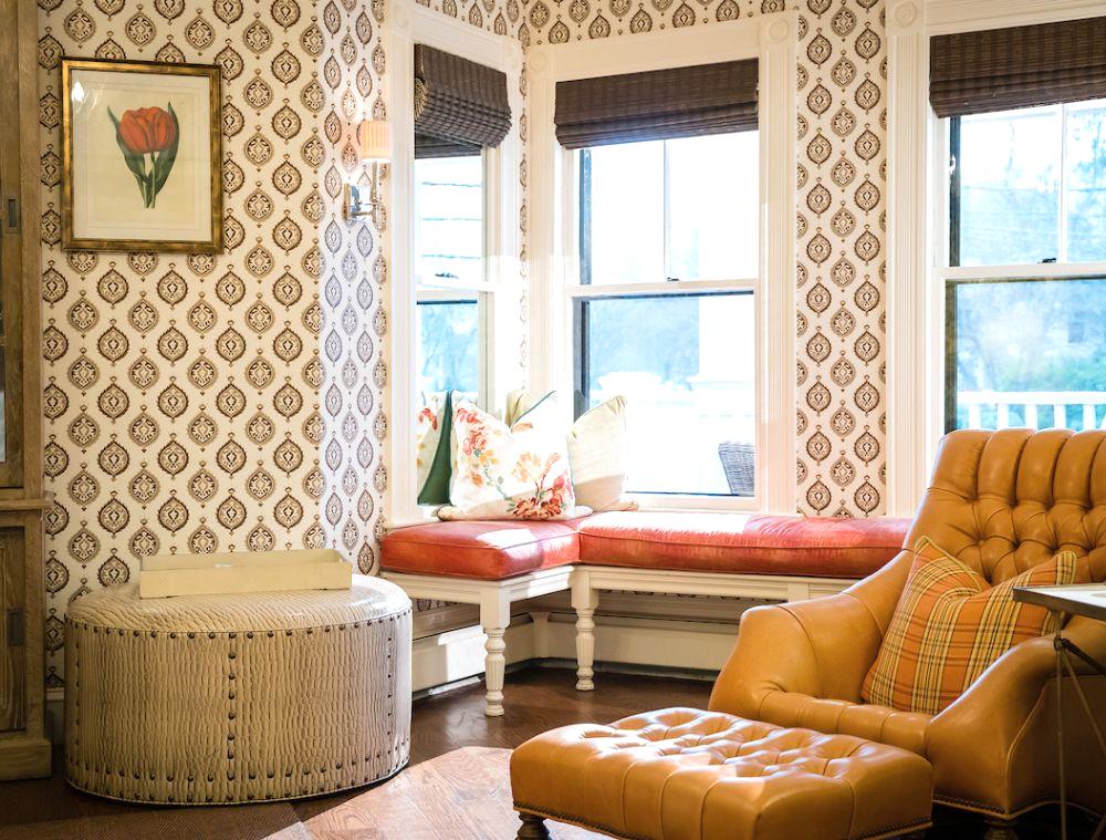 Cozy living room with patterned wallpaper, orange leather armchair, tufted ottoman, window bench with red cushion, and floral pillows by large windows.