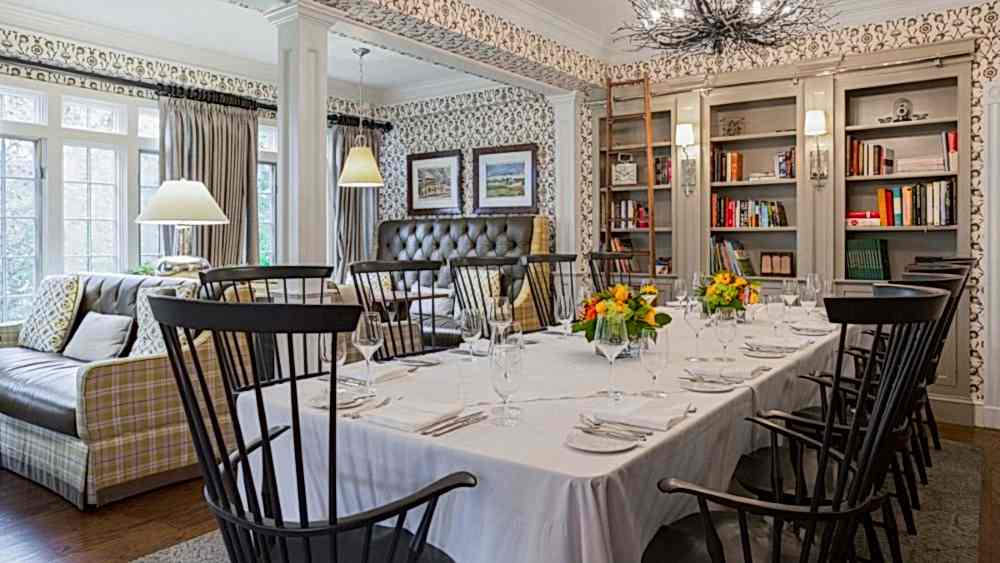Elegant dining room with a long table set for a meal, surrounded by black chairs, bookshelves, a sofa, and large windows letting in natural light.