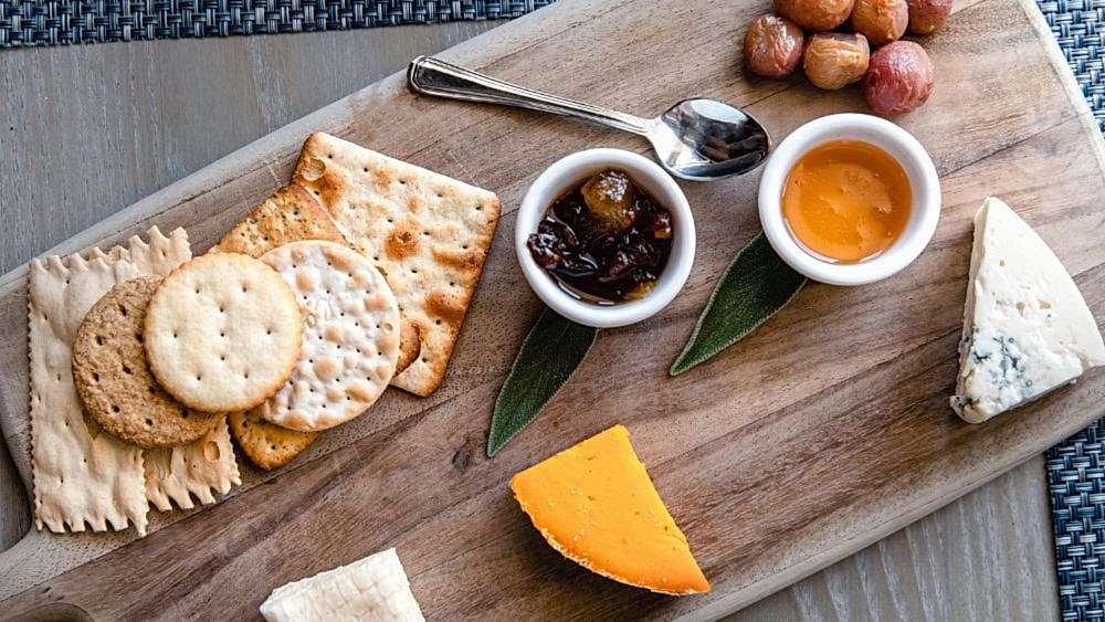 A cheese board with assorted crackers, cheddar and blue cheese, honey, chutney, nuts, and a spoon on a wooden serving board.