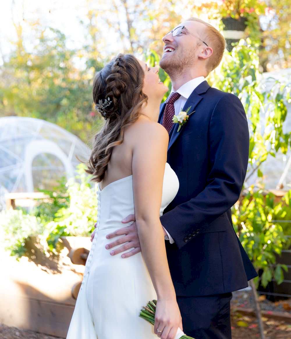 A bride and groom stand outdoors, smiling and laughing together, surrounded by greenery and sunlight.