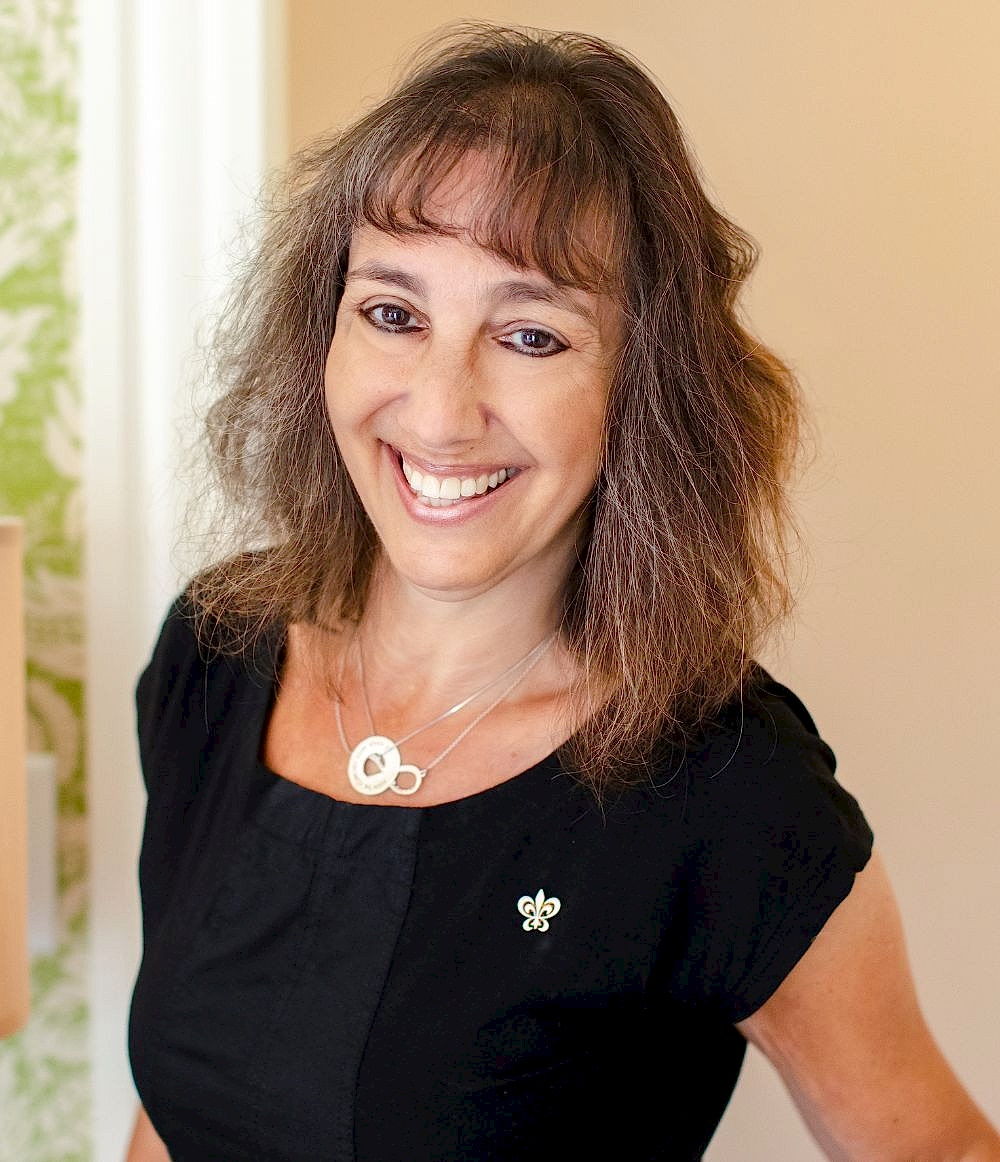 Woman with wavy brown hair, wearing a black top and smiling, stands indoors. She has a silver necklace and a fleur-de-lis pin on her shirt.