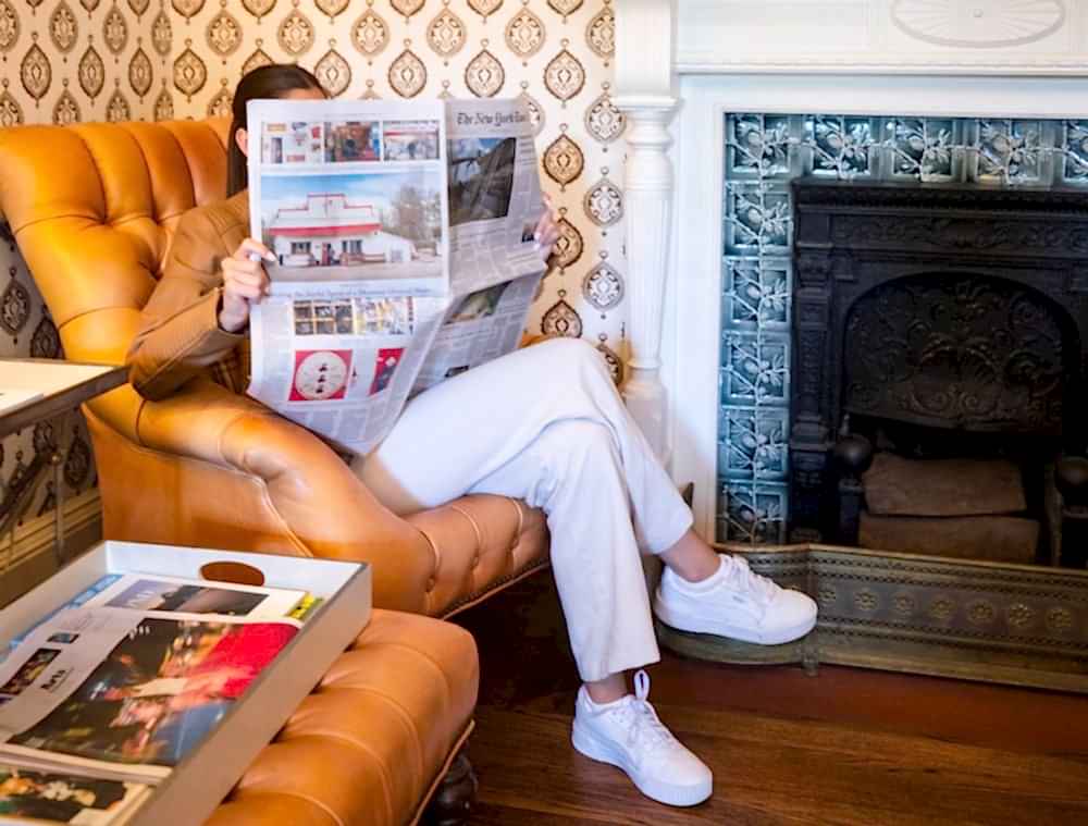 A person sits in a tan armchair reading a newspaper, with a tray of magazines nearby and a decorative fireplace in the background.