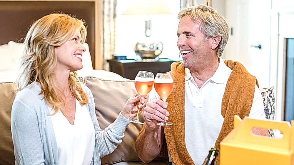 Smiling middle-aged couple sitting on a couch, clinking glasses of rosé wine and looking at each other happily.
