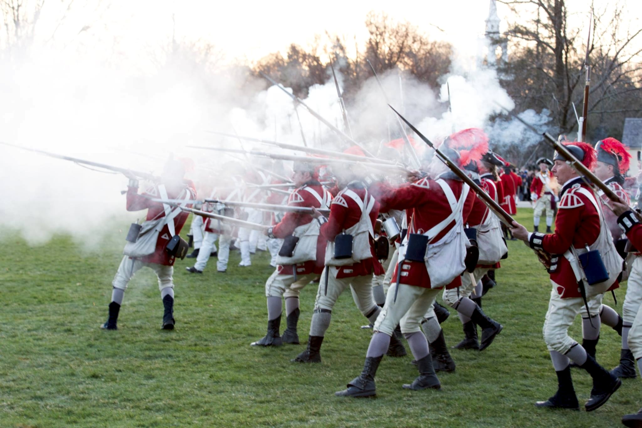 Historical reenactors in red coats fire muskets, creating clouds of smoke, while advancing on a grassy field during a battle reenactment.