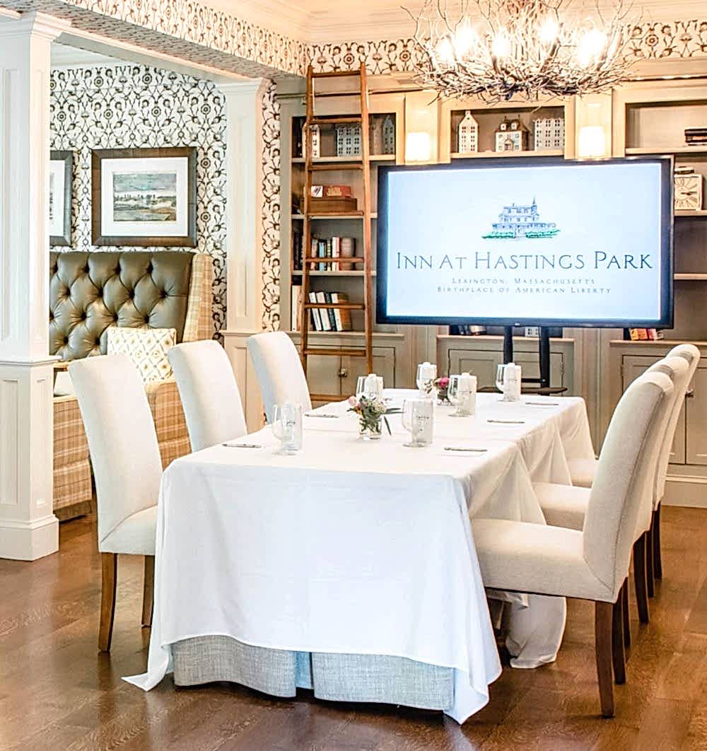 Elegant dining room with six white chairs around a table, set for a meal, and a large screen displaying Inn at Hastings Park.