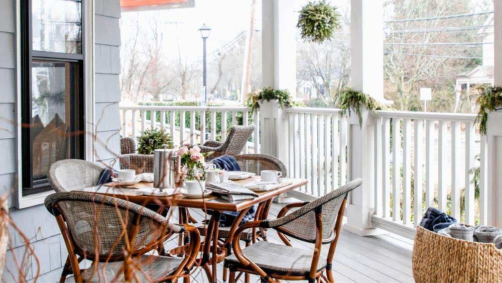 A cozy porch with a wooden table set for breakfast, surrounded by wicker chairs, greenery, and white railing overlooking a quiet street.