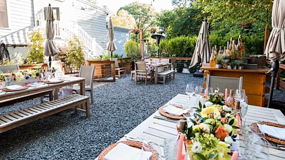 Outdoor dining area with wooden tables set for a meal, floral centerpieces, benches, umbrellas, and lush greenery in the background.