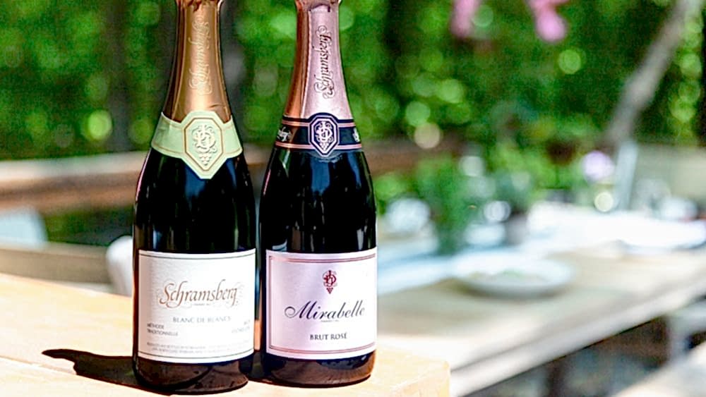 Two bottles of sparkling wine, Schramsberg Blanc de Blancs and Mirabelle Brut Rosé, on a wooden table outdoors with greenery in the background.