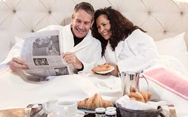 A smiling couple in white robes enjoys breakfast and reads a newspaper together in bed, with croissants, coffee, and jam on a tray in front of them.