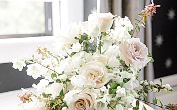 A bouquet of pale pink and white roses mixed with white flowers and greenery, set near a sunlit window indoors.
