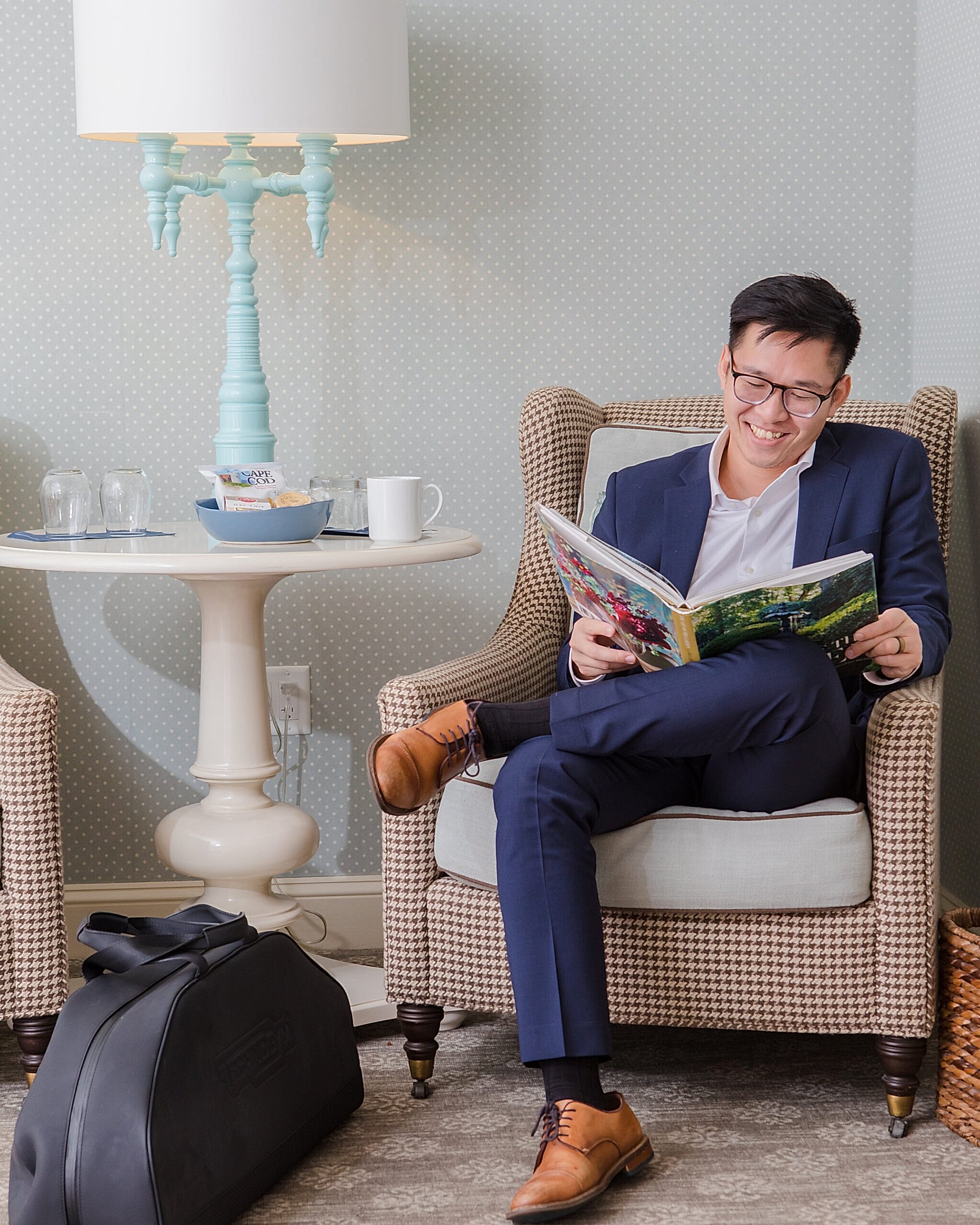 A smiling man in a suit sits in a chair reading a magazine, with a travel bag on the floor and a side table with a lamp, mug, and snacks nearby.