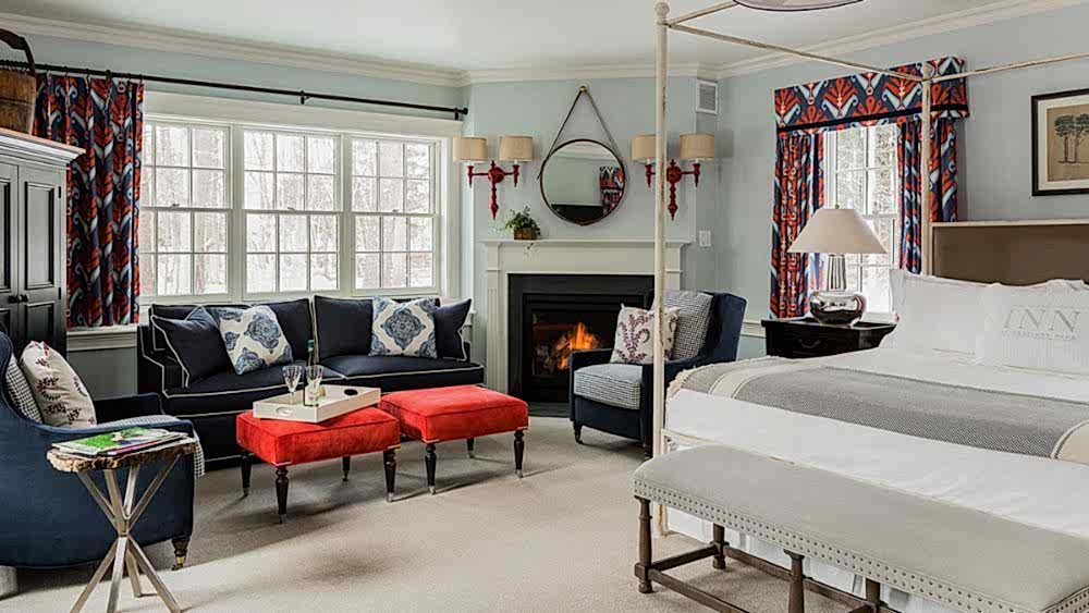 Bright, elegant bedroom with a four-poster bed, navy sofas, red ottomans, a fireplace, and large windows with patterned curtains.
