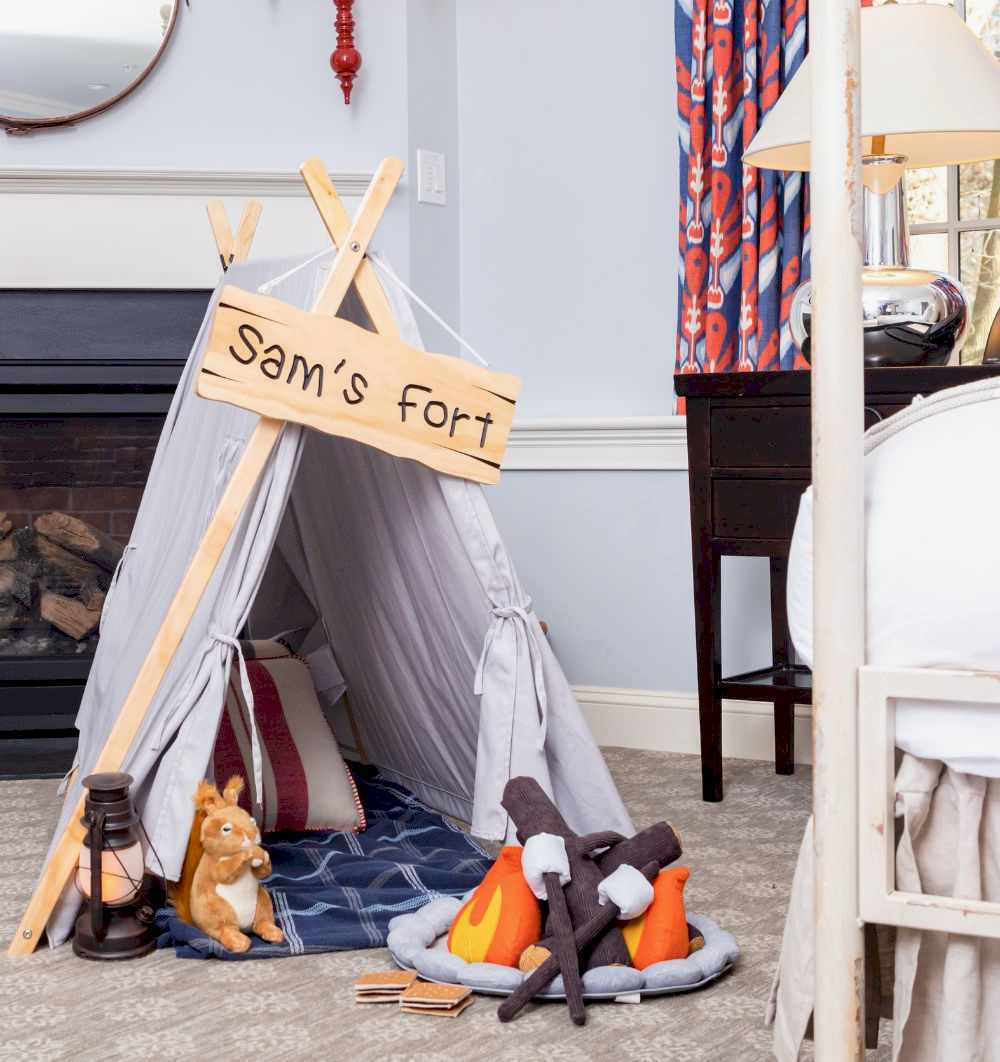 A childrens play tent labeled Sams fort with plush campfire, lantern, and toy kangaroo set up indoors next to a bed and desk.