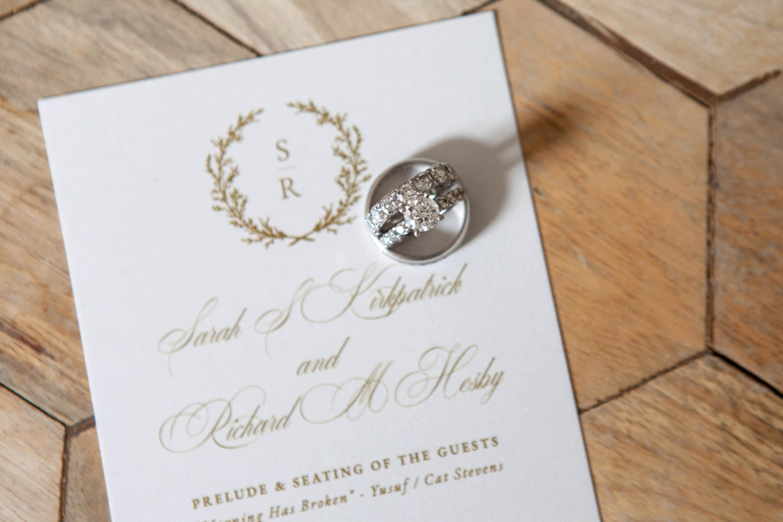 A wedding invitation with rings on top, displaying the names Sarah Kirkpatrick and Richard M. Hosby on a wooden surface.