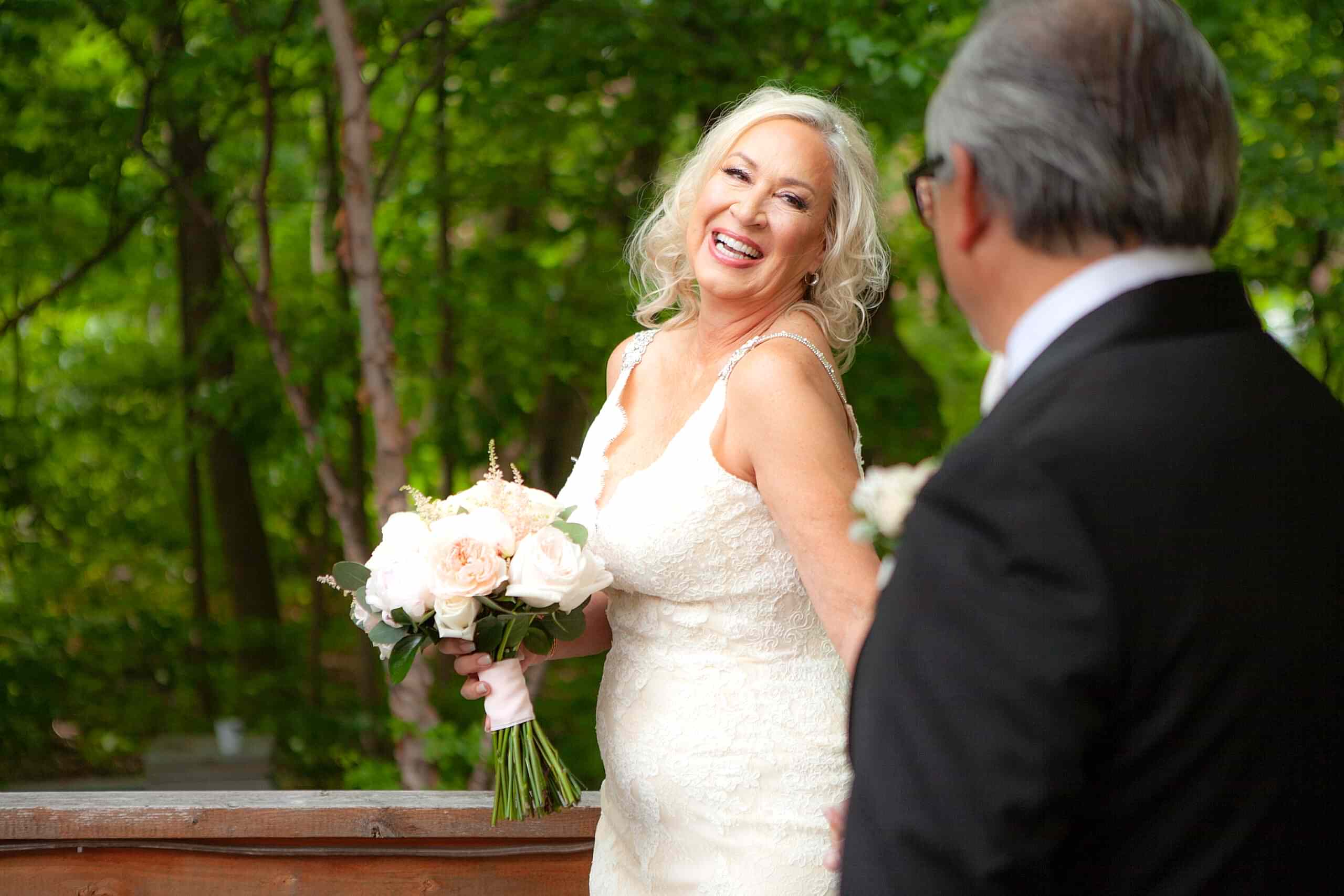 A joyful bride in a white dress holding a bouquet smiles at a man in a suit outdoors with green trees in the background.