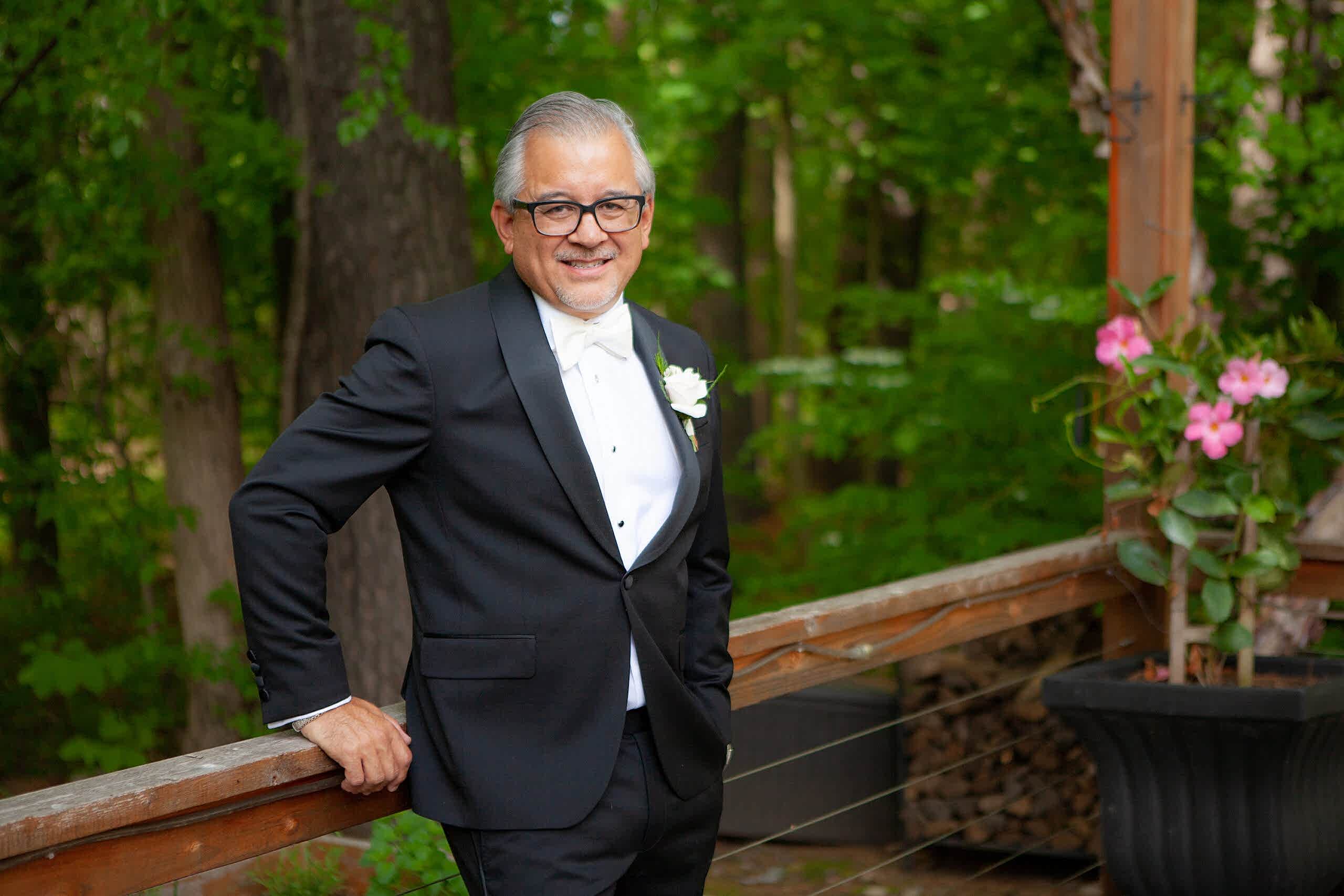 An older man in a black tuxedo and white bow tie smiles while leaning on a wooden railing outdoors, with green trees and potted flowers behind him.