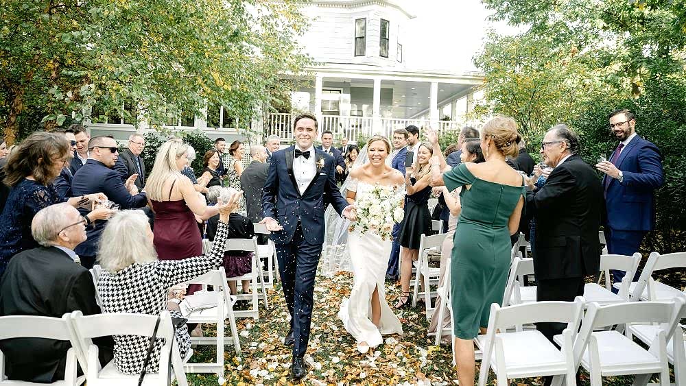 A bride and groom smile as they walk down the aisle outdoors, surrounded by cheering guests and autumn leaves.