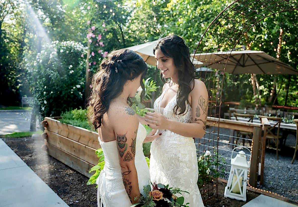 Two brides in white dresses smile at each other outdoors, surrounded by greenery and sunlight, during their wedding ceremony.
