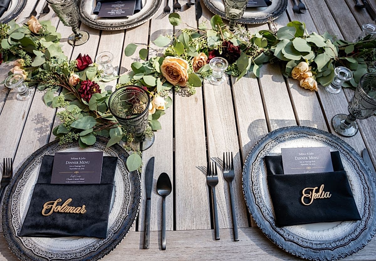 Elegant outdoor table setting with floral garland, ornate plates, menus, and black napkins embroidered with the names Telma and Golmar.