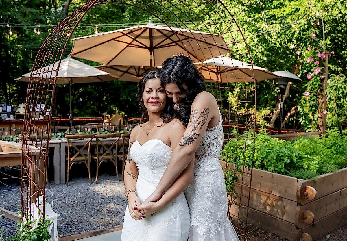 Two brides in white dresses embrace and smile in an outdoor garden setting decorated with umbrellas and wooden tables.